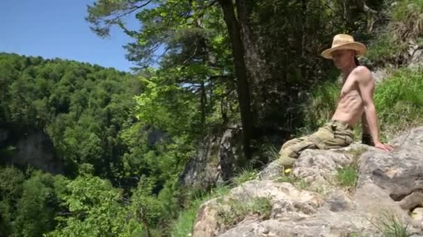 Man med naken överkropp sitter på toppen klippa, tittar ner. Sommar, solig dag, panoramautsikt — Stockvideo