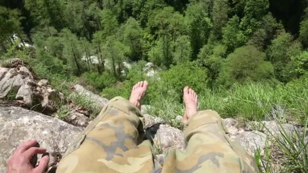 Mannen i militärbyxor sitter på klippan och ser ner på ravinen. Första person, närbild — Stockvideo