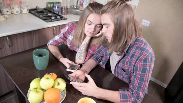 Een jong paar mellenialen met lang haar zitten aan de keukentafel met telefoons in hun handen. Surfen en winkelen op het internet vanaf mobiele apparaten. Begrip modern jong gezin — Stockvideo