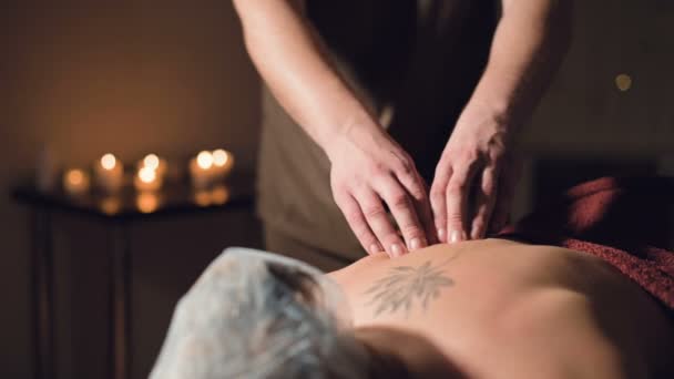 Ung manlig massage terapeut gör rygg massage till en kvinna med en tatuering i ett massagerum med svagt ljus på bakgrunden av ljus. Låg nyckel premium massage koncept — Stockvideo