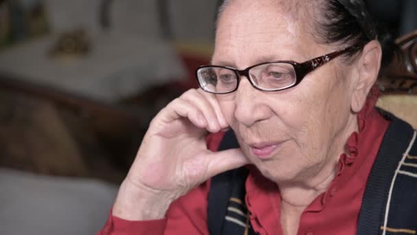 Портрет літньої жінки-пенсіонера в окулярах, що спирається на її руку в приміщенні, сидячи і думаючи. Літня жінка 80 років — стокове відео