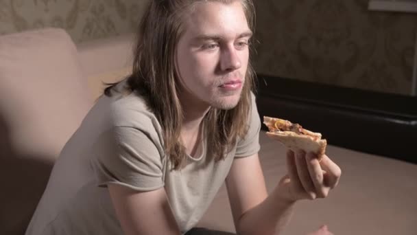 Unge man sitter i vardagsrummet på soffkvällen, tittar på TV, äter pizza. Närbild — Stockvideo