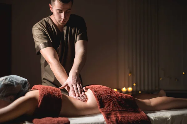 Professional premium massage in a dark atmospheric cabinet. Young man doing massage to a female client in a dark office on the background of burning candles