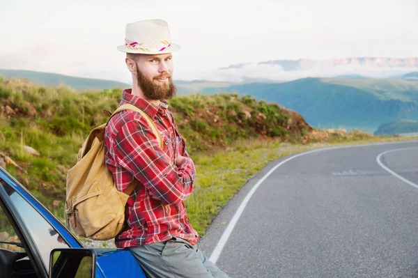 Retrato de um viajante sério feliz barbudo hipster com uma mochila em uma camisa xadrez e um chapéu ao lado de um carro desconhecido fica na estrada ao pôr do sol nas montanhas. Conceito de viagem feliz e confiante — Fotografia de Stock