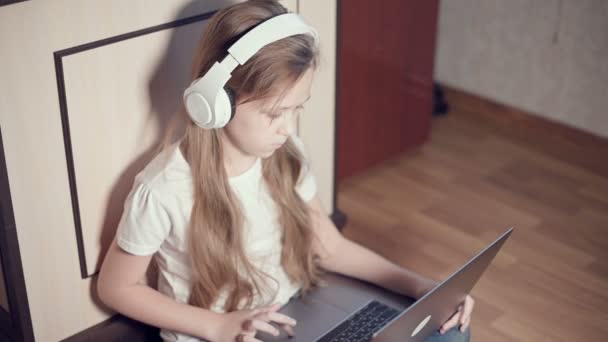 Une petite fille intelligente de sept ans en casque blanc avec un ordinateur portable dans les mains pousse sur le sol dans sa chambre. La jeune génération sur Internet et les technologies de l'information — Video