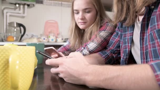 In Großaufnahme sitzt ein junges Paar Mellenials mit langen Haaren am Küchentisch, Telefone in der Hand. Surfen und Einkaufen im Internet von mobilen Geräten aus. Konzept der modernen jungen Familie — Stockvideo