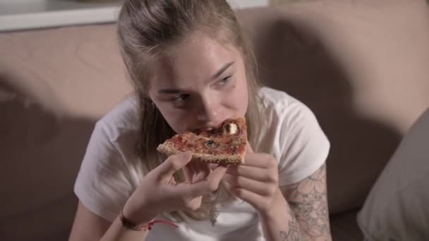 Jeune femme mignonne est assise dans le salon sur le canapé, regarde la télévision, mange de la pizza. Gros plan — Video