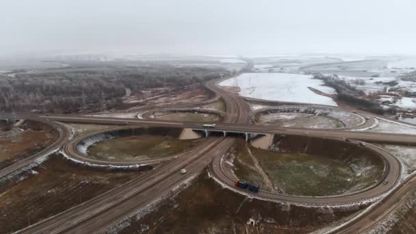 Flygfoto av bilar och lastbilar som rör sig i en vinterkorsning är en stor vägkorsning. Obemannad utsikt över biltrafiken på en motorväg i förorterna på vintern. Rondellen vid korsningen — Stockvideo