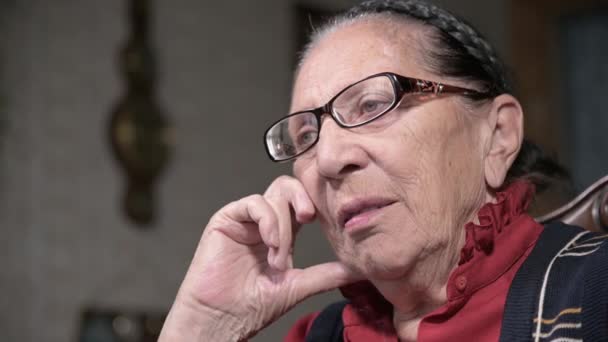 Портрет пожилой задумчивой женщины в очках, опирающейся на руку в помещении, сидящей и думающей. Пожилая женщина 80 лет — стоковое видео