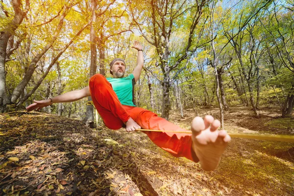 Amplo ângulo masculino tightrope walker balanceamento enquanto sentado descalço no slackline na floresta de outono. O conceito de esportes ao ar livre e vida ativa de pessoas idosas — Fotografia de Stock