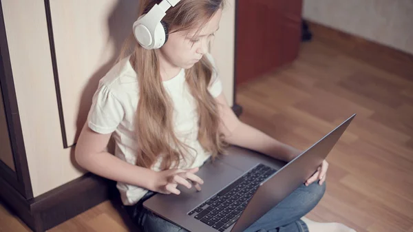 Yedi yaşında, elinde dizüstü bilgisayarla beyaz kulaklıklı akıllı bir kız odasında yere bastırıyor. İnternet ve bilişim teknolojisindeki genç nesil. — Stok fotoğraf