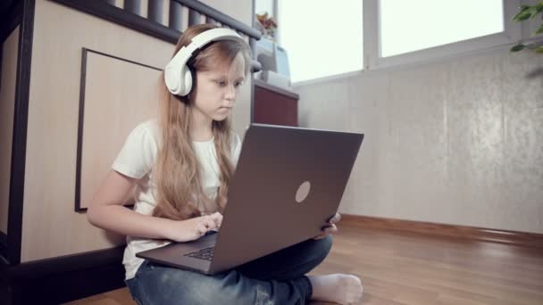 Una niña inteligente de siete años con auriculares blancos con un portátil en las manos está empujando al suelo en su habitación. La generación joven en Internet y la tecnología informática — Vídeos de Stock