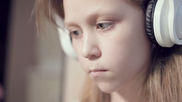 Um close-up de um retrato plano apressado de uma menina séria pensativa e destacada que tem 10 anos de idade em grandes fones de ouvido brancos dentro de casa. Olho lateral — Vídeo de Stock