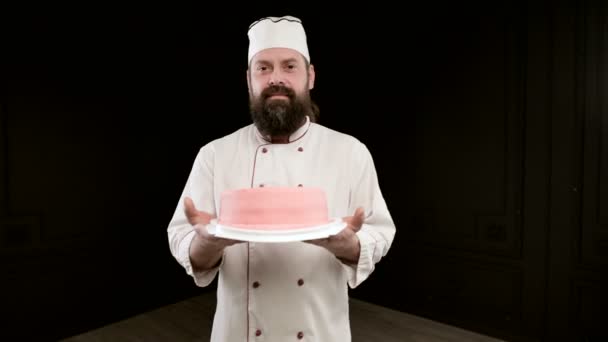Homem cozinhe em uniforme com barba no fundo preto, mantém o bolo nas mãos. Fechar. — Vídeo de Stock