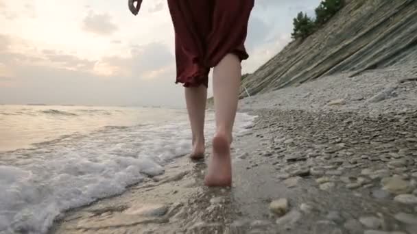 Vista trasera de cerca de las piernas Chica caucásica camina a lo largo del agua en la costa de piedra del mar hacia el sol poniente al atardecer con olas — Vídeos de Stock