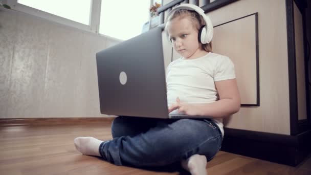 Una niña inteligente de siete años con auriculares blancos con un portátil en las manos está empujando al suelo en su habitación. La generación joven en Internet y la tecnología informática — Vídeo de stock