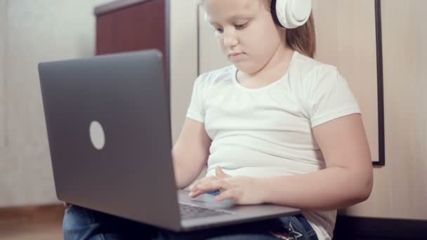 一个7岁的聪明小女孩，手里拿着笔记本电脑，戴着白色耳机，在她房间的地板上推挤着。在互联网和IT技术上的年轻一代 — 图库视频影像