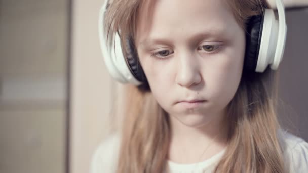 Eine Nahaufnahme eines überstürzten Planporträts eines ernsthaft nachdenklichen und distanzierten Mädchens, das 10 Jahre alt ist und große weiße Kopfhörer trägt. Seitenblick — Stockvideo