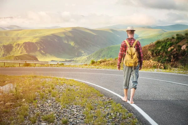 Stílusos mezítláb szakállas férfi stoppal utazó egy kalap és egy hátizsák sétál végig egy ország út a hegyekben naplementekor. A kilátás a hátsó. Utazási koncepció pénz nélkül. — Stock Fotó