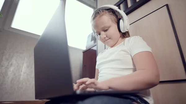 Een slim meisje van zeven met een witte koptelefoon met een laptop in haar handen duwt op de vloer in haar kamer. De jonge generatie op het internet en IT-technologie — Stockfoto