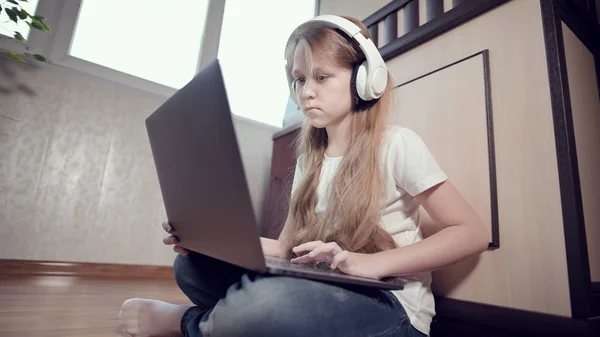 Een slim meisje van zeven met een witte koptelefoon met een laptop in haar handen duwt op de vloer in haar kamer. De jonge generatie op het internet en IT-technologie — Stockfoto