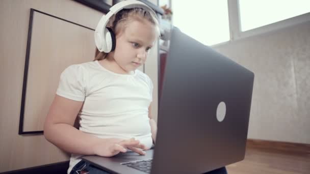 Uma menina esperta de sete anos de idade em fones de ouvido brancos com um laptop em suas mãos está empurrando no chão em seu quarto. A geração jovem na Internet e tecnologia de TI — Vídeo de Stock