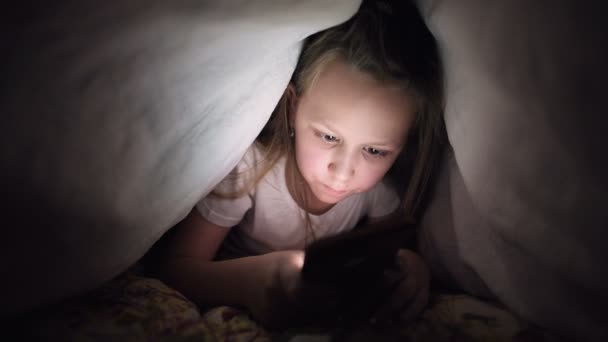 A young little girl is hiding under a blanket to use a digital tablet smartphone device late after sleep. Loneliness of young children and rescue on the phone. — 비디오