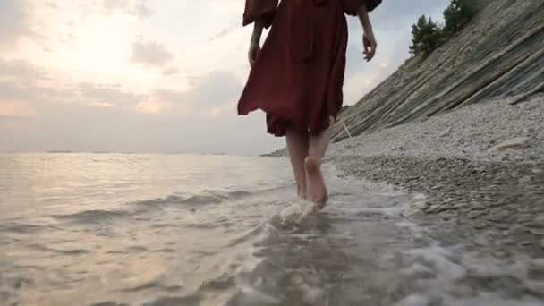 Close-up rear view of legs Caucasian girl walks along the water on the stone coast of the sea towards the setting sun at sunset with waves — Stock Video