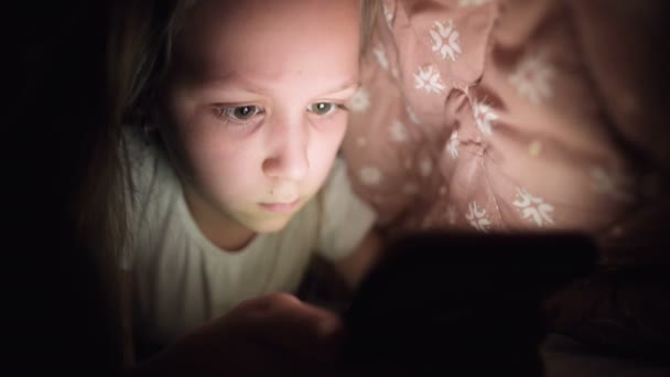 Una giovane bambina si nasconde sotto una coperta per utilizzare un dispositivo smartphone tablet digitale in ritardo dopo il sonno. Solitudine dei bambini piccoli e salvataggio al telefono . — Video Stock