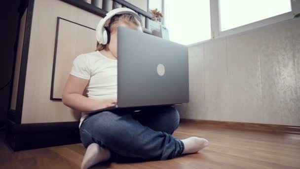 Una niña inteligente de siete años con auriculares blancos con un portátil en las manos está empujando al suelo en su habitación. La generación joven en Internet y la tecnología informática — Vídeo de stock