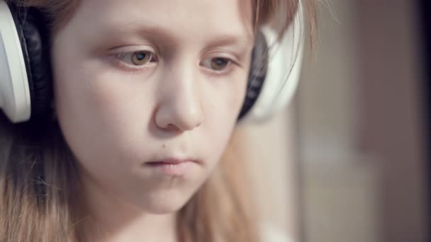 Um close-up de um retrato plano apressado de uma menina séria pensativa e destacada que tem 10 anos de idade em grandes fones de ouvido brancos dentro de casa. Olho lateral — Vídeo de Stock