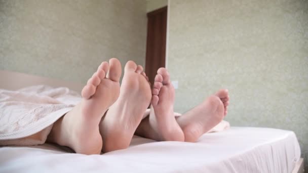 Nahaufnahme der Füße eines jungen Paares, das unter den Abdeckungen im Schlafzimmer hervorsteht. Barfüßige streicheln einander bei der Körperpflege. — Stockvideo