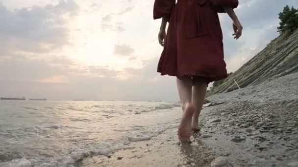 Close-up rear view of legs Caucasian girl walks along the water on the stone coast of the sea towards the setting sun at sunset with waves — Stock Video