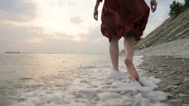 Kafkasyalı kız, gün batımında güneşin batışına doğru denizin taştan kıyısı boyunca dalgalarla yürür.