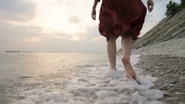 Close-up rear view of legs Caucasian girl walks along the water on the stone coast of the sea towards the setting sun at sunset with waves — Stock Video