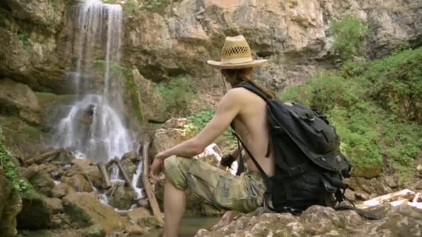 El hombre con el torso desnudo se sienta en piedra sobre el fondo de la cascada. Primer plano — Vídeo de stock