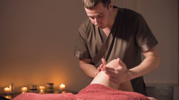 O tiro médio de um homem massagista profissional faz com que um cliente massageie as pernas e joelhos em um escritório com iluminação fraca contra o fundo de velas acesas. Serviço de massagem premium para clientes de luxo — Vídeo de Stock