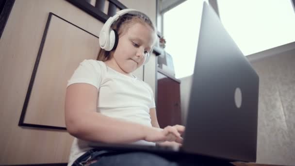 Une petite fille intelligente de sept ans en casque blanc avec un ordinateur portable dans les mains pousse sur le sol dans sa chambre. La jeune génération sur Internet et les technologies de l'information — Video