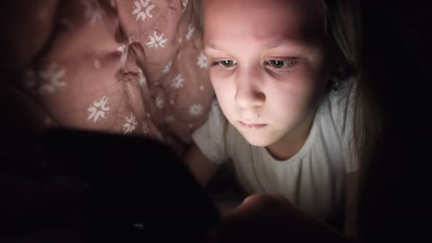 A young little girl is hiding under a blanket to use a digital tablet smartphone device late after sleep. Loneliness of young children and rescue on the phone. — Stock Video