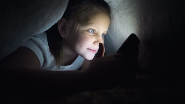 Una niña pequeña se esconde debajo de una manta para usar un dispositivo de teléfono inteligente tableta digital tarde después de dormir. Soledad de los niños pequeños y rescate por teléfono . — Vídeos de Stock