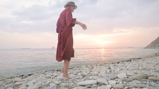 Close-up slow motion following behind the legs of a barefoot girl in a red dress fluttering in the wind at sunset walking crouching along the stones of the sea shore. Light Moments of Tidelessness — Stock Video