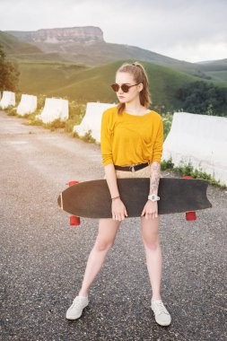 Güneş gözlüğü güzel ve şık genç kadın ve bir dövme ile günbatımı gökyüzüne karşı bir kaykay veya longboard ile pozlar