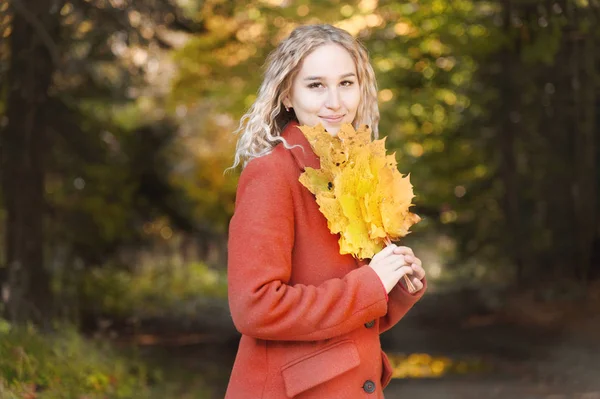 Vonzó fehér fiatal szőke lány portréja piros kabátban, egy csokor lehullott sárga levéllel, ami az őszi erdőben mosolyog. Az őszi és őszi ünnepek és hétvégék fogalma — Stock Fotó