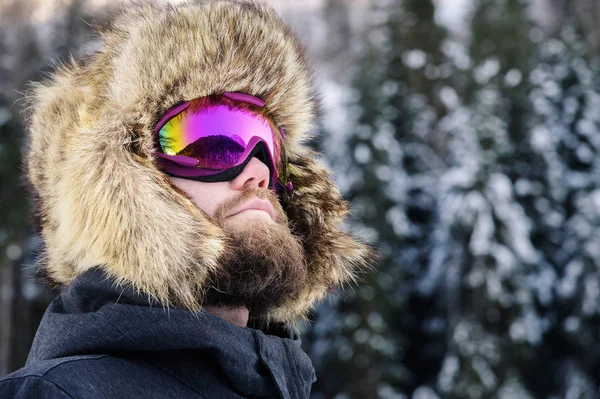 Retrato fechado de um esquiador de snowboard feliz barbudo em uma máscara de esqui com óculos e um chapéu de velha escola grande de pele em um contexto de umas montanhas nevadas de inverno — Fotografia de Stock