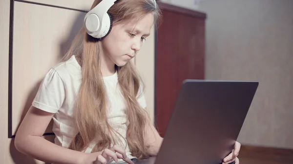 Een slim meisje van zeven met een witte koptelefoon met een laptop in haar handen duwt op de vloer in haar kamer. De jonge generatie op het internet en IT-technologie — Stockfoto