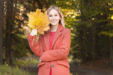 Sonbahar ormanında gülümseyen sarı yapraklarla dolu bir buketle kırmızı paltolu çekici sarışın kızın portresi. Sonbahar, sonbahar tatilleri ve hafta sonları konsepti.