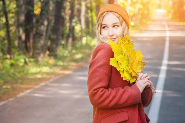 Portret atrakcyjnej białej blondynki w czerwonym płaszczu w czerwonym kapeluszu z bukietem opadłych żółtych liści uśmiechających się w jesiennym lesie i przykrywających jej twarz liśćmi. — Zdjęcie stockowe