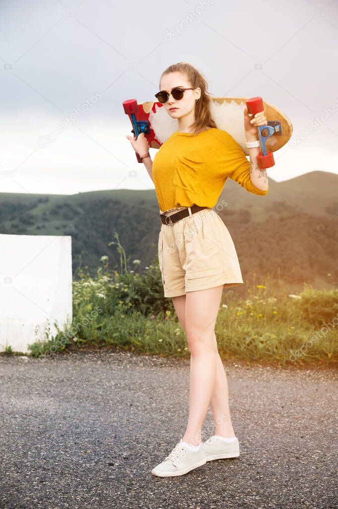 Beautiful and fashionable young woman in sunglasses and with a tattoo poses with a skateboard or longboard against the sunset sky