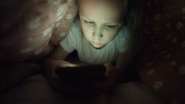 Молода маленька дівчинка ховається під ковдрою, щоб використовувати цифровий планшетний пристрій смартфона пізно після сну. Самотність маленьких дітей і порятунок по телефону . — стокове відео