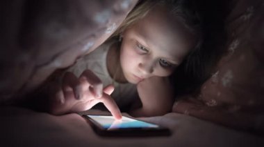 Küçük bir kız uyuduktan sonra dijital tablet akıllı telefon cihazı kullanmak için battaniyenin altına saklanıyor. Küçük çocukların yalnızlığı ve telefonda kurtarma..
